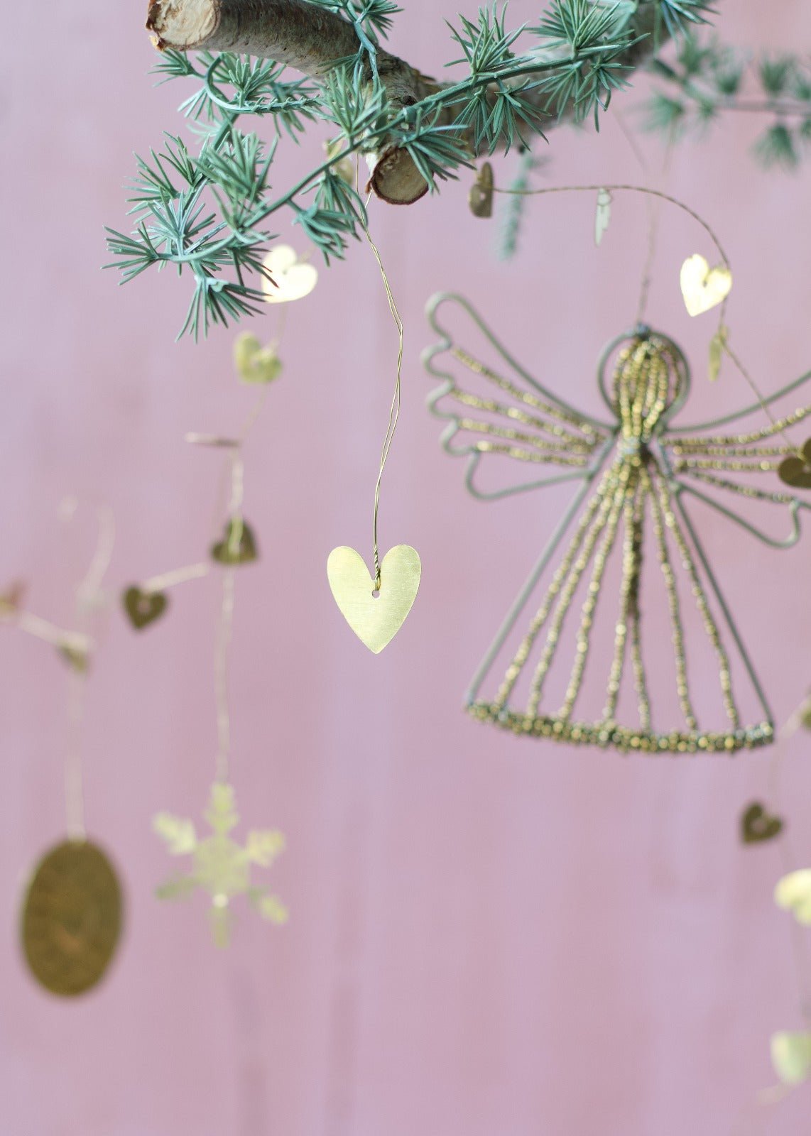 Brass heart garland hanging from pine branch with a pink background, featuring small golden hearts and a decorative wire angel.