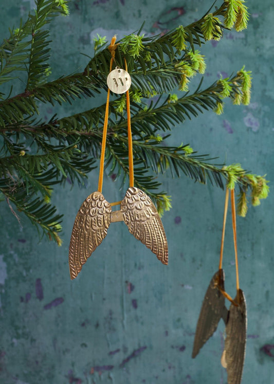 Large golden brass angel wings ornament with embossed feather details, hanging on a green fir branch.