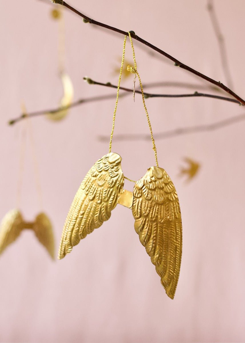 Small gold brass angel wings ornament hanging from a branch against a soft pink background