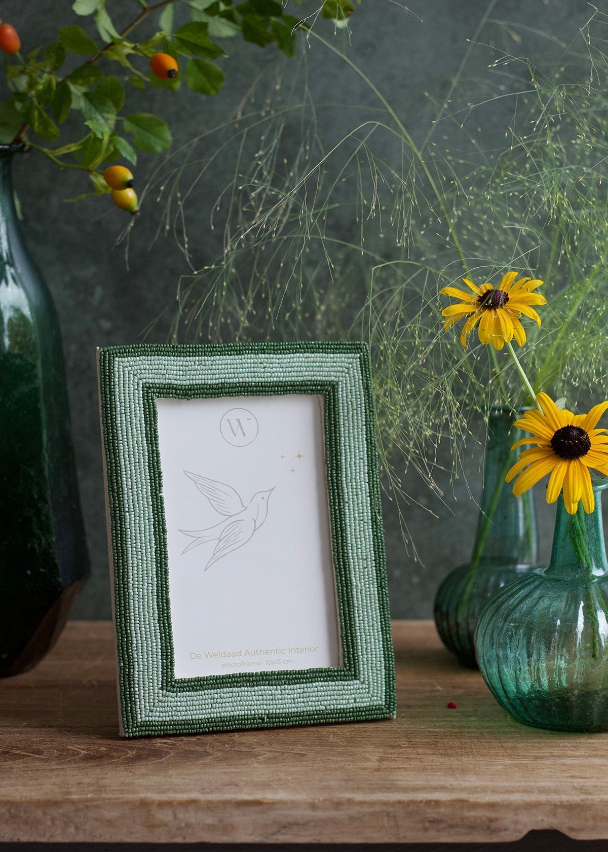 Green framed picture on a wooden surface with yellow flowers and green vases in the background.
