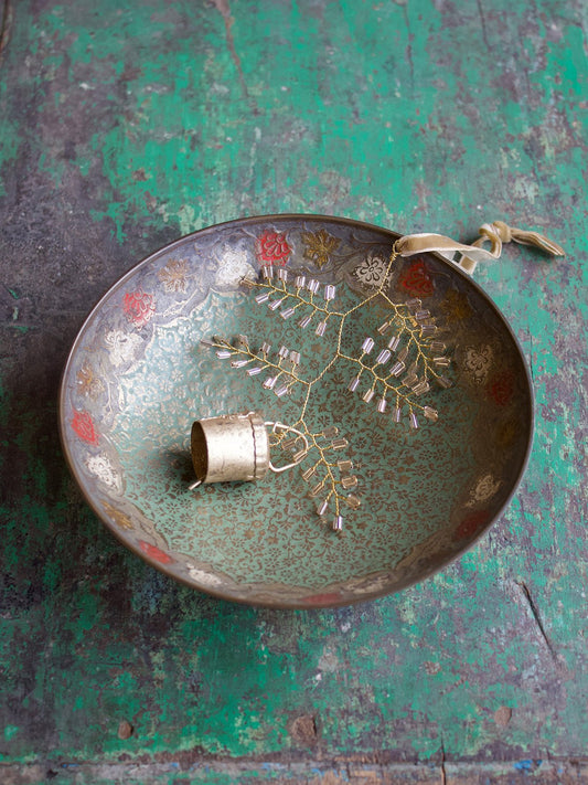 Decorative vintage metal bowl with floral patterns, holding a delicate champagne-coloured beaded branch ornament and a small brass bell, placed on a weathered green wooden surface.