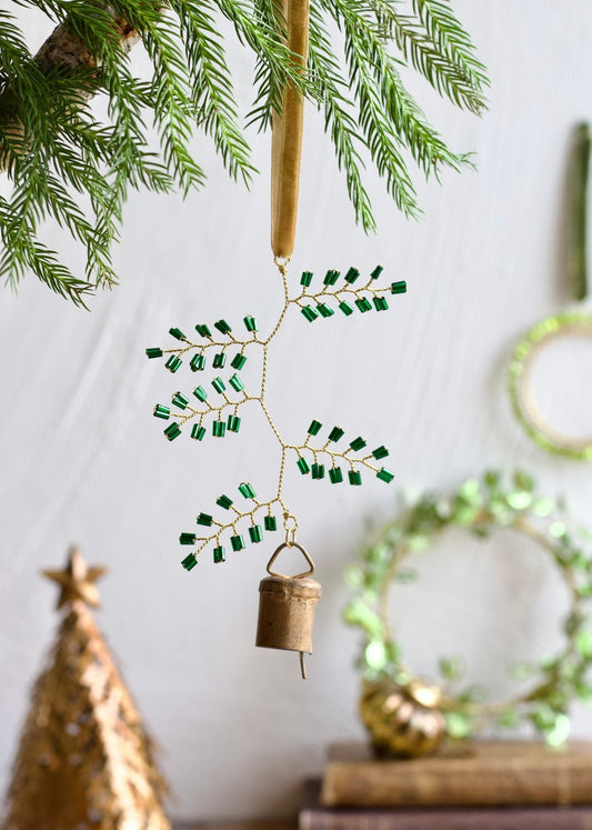 Handmade green beaded ornament with brass bell hanging from a golden velvet ribbon on a pine branch, with festive decor in the background.