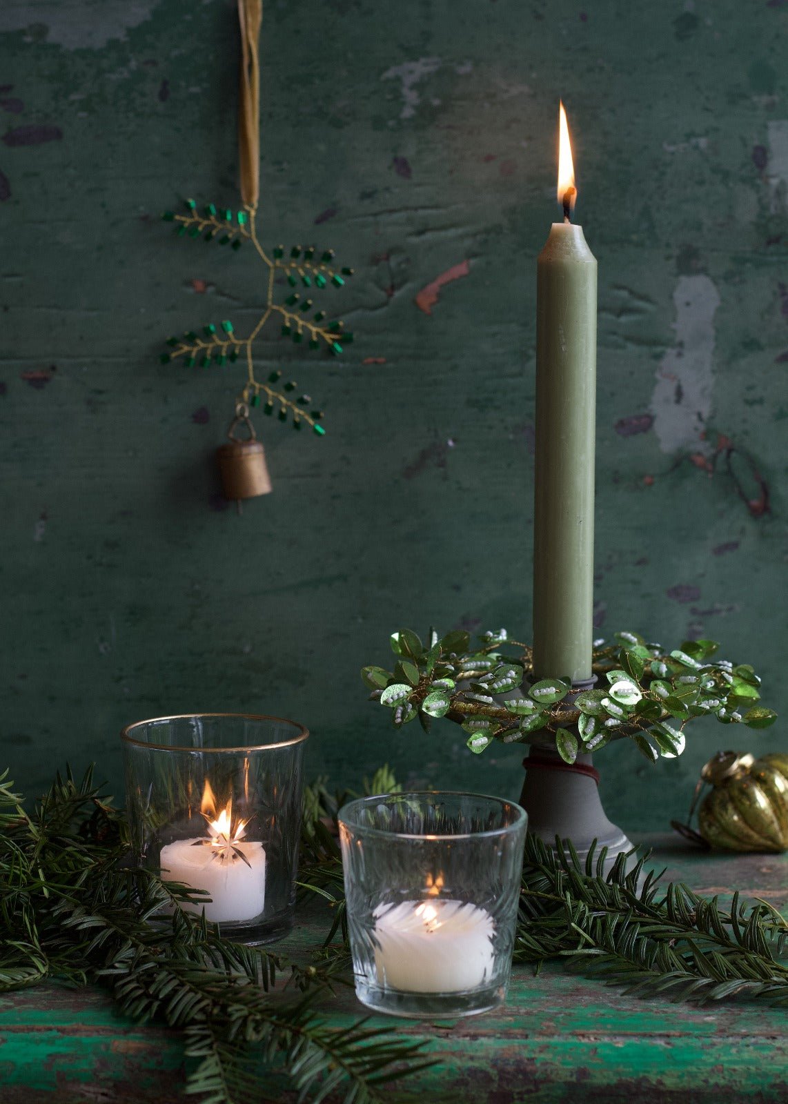 A green candle in a candlestick decorated with glass leaves, surrounded by pine branches and tealight holders, with a beaded green ornament and bell hanging on a velvet ribbon in the background.