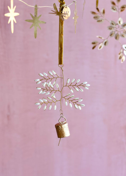 Brass ornament with delicate white beads arranged in a snowflake shape, hanging from a golden velvet ribbon with a small vintage-style bell attached at the bottom.
