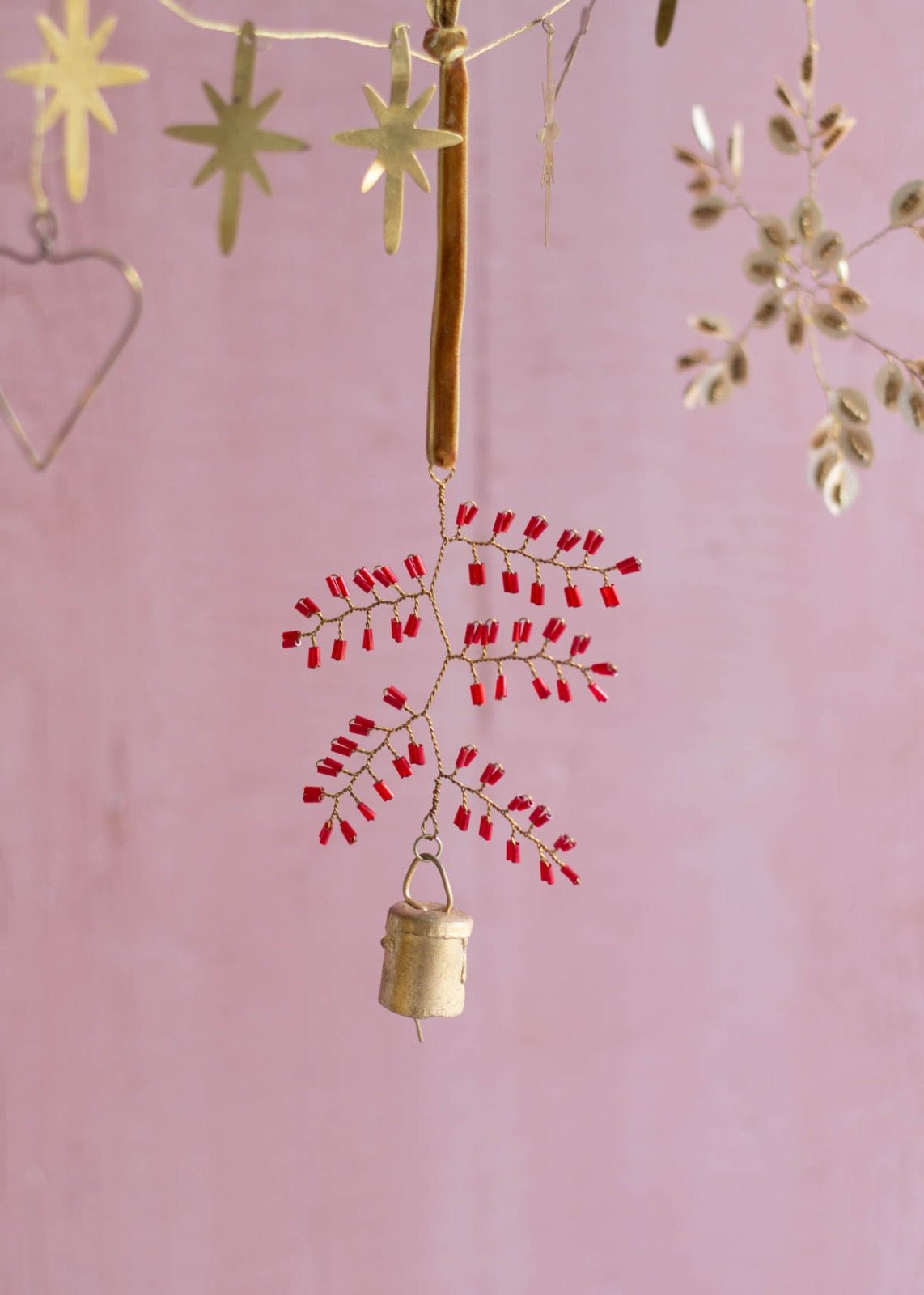 Handmade brass snowflake ornament with red glass beads and a small bell, hanging from a golden velvet ribbon against a soft pink background.