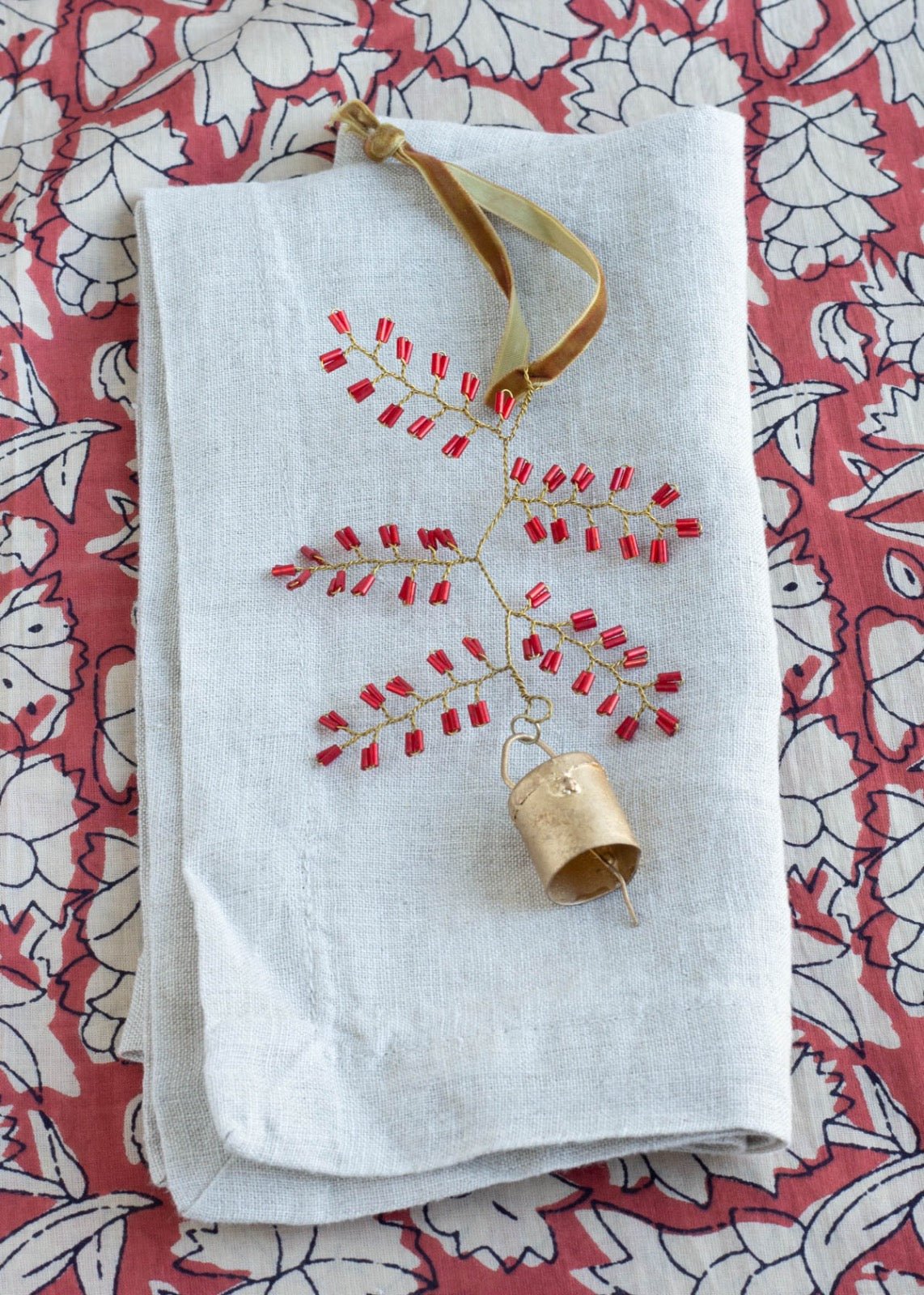 Handmade ornament with red glass beads and brass bell, displayed on folded linen fabric with a floral patterned background.