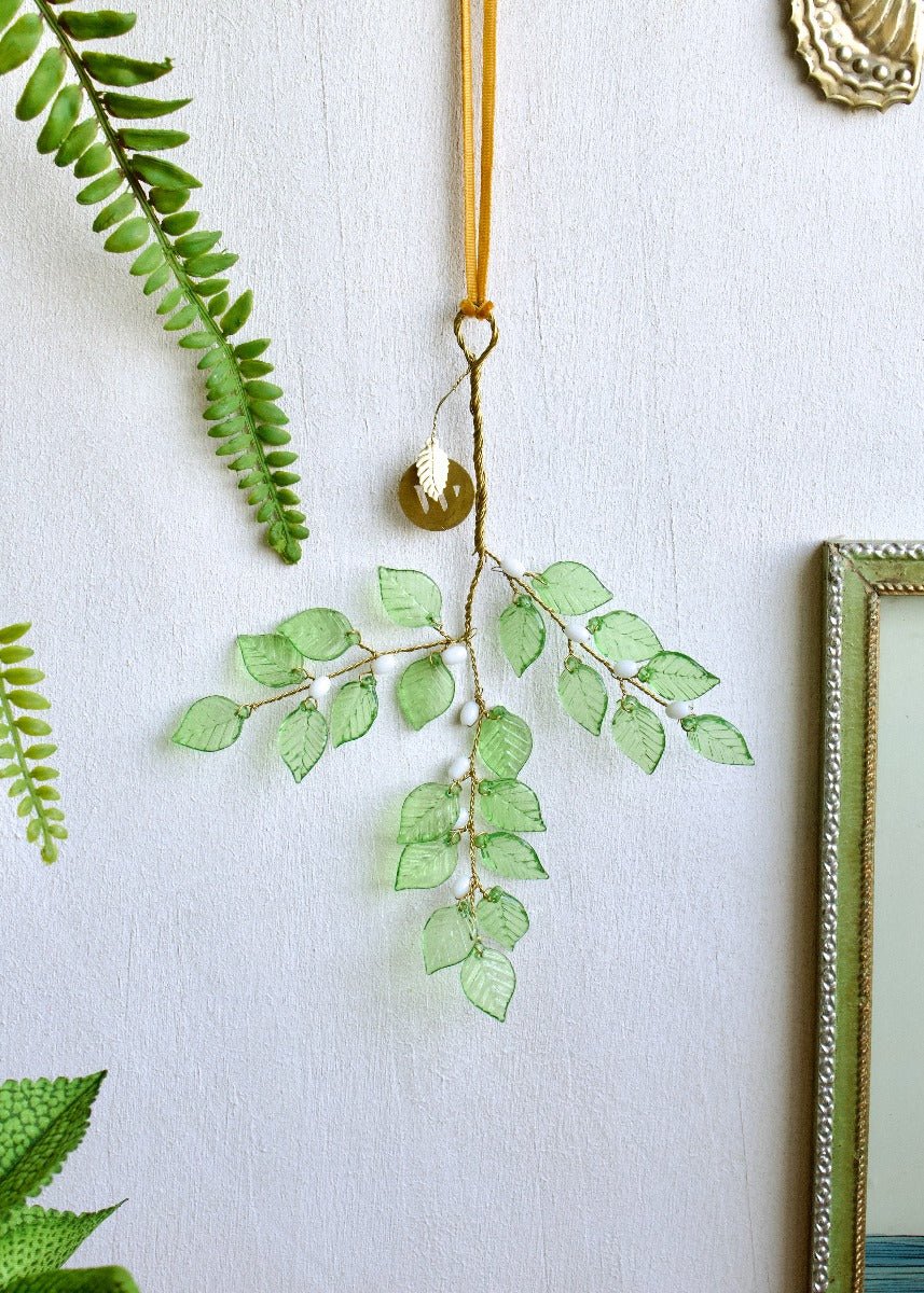 Decorative hanging branch with translucent green leaves and white bead details, shown on a white wall.
