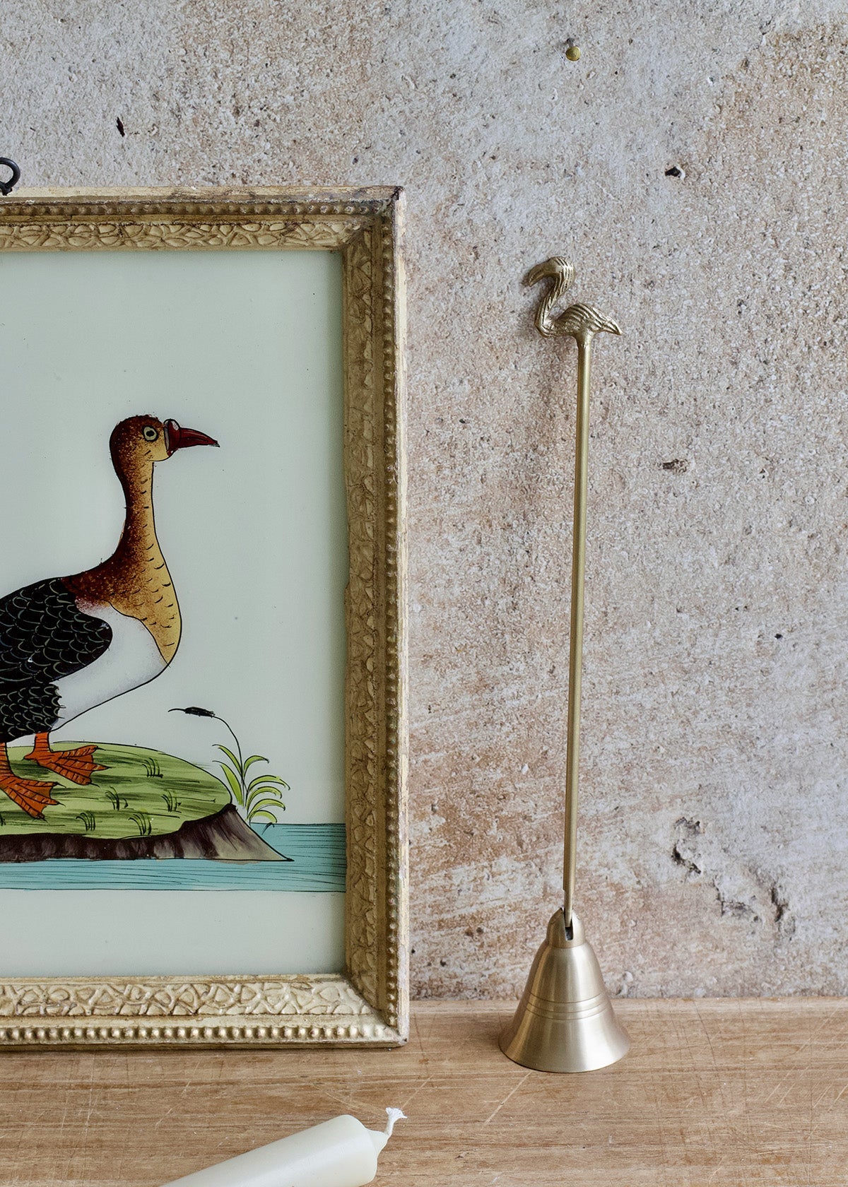 Brass candle snuffer next to a framed illustration of a duck on a textured wall.
