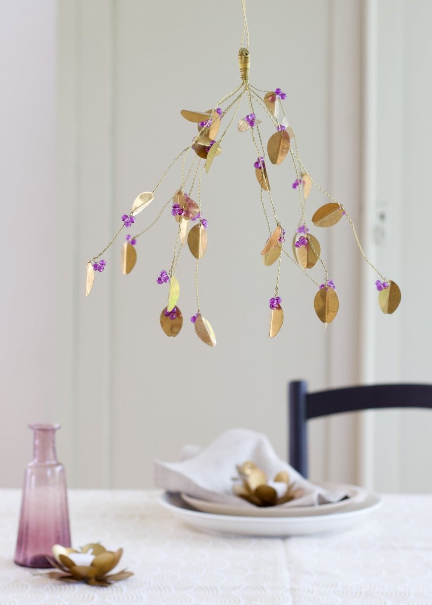 Small decorative brass bunch with oval leaves and purple beads, hanging above a table set with a soft linen napkin and vintage pink vase.