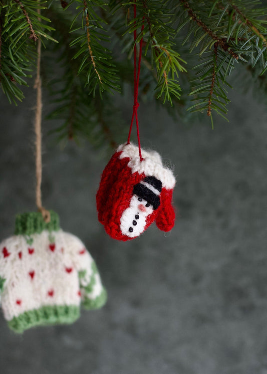 Knitted Christmas ornaments, one in a red sweater with a snowman design, the other in a green sweater with red hearts, hanging from a tree.