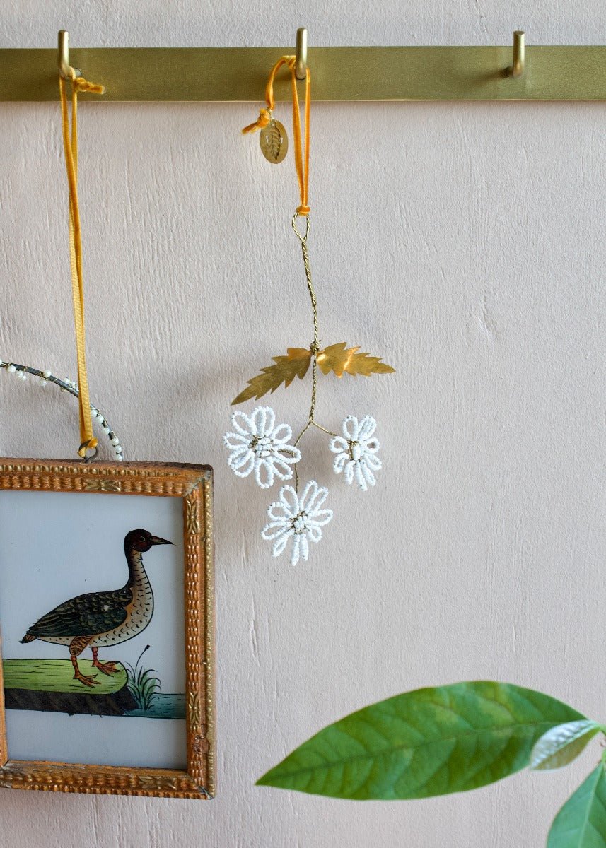 Beaded flower ornament with brass leaves and ochre ribbon hanging on brass hook.