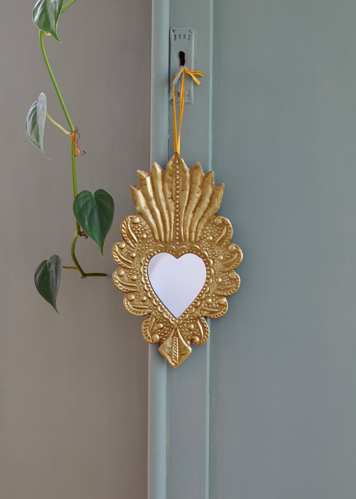 Decorative gold heart-shaped mirror hanging on a light blue door with a green plant in the background.