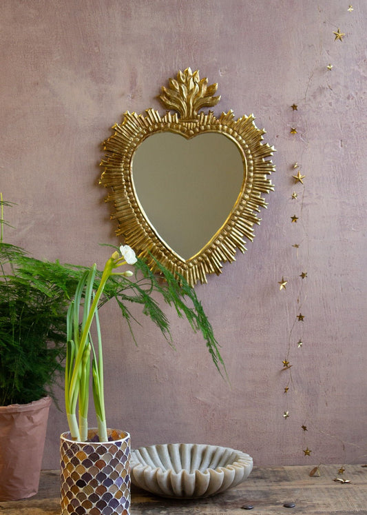 Ex-voto heart-shaped mirror with golden rays and flame crown on a pink wall.