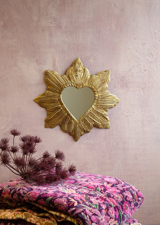 Handcrafted heart-shaped brass wall mirror with a hammered leaf design and small cross detail, displayed against a soft pink plastered wall with folded floral quilts in the foreground.