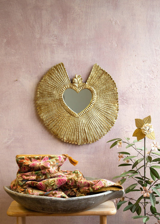 Ex-voto heart mirror with wide radiant golden frame on a pink wall.
