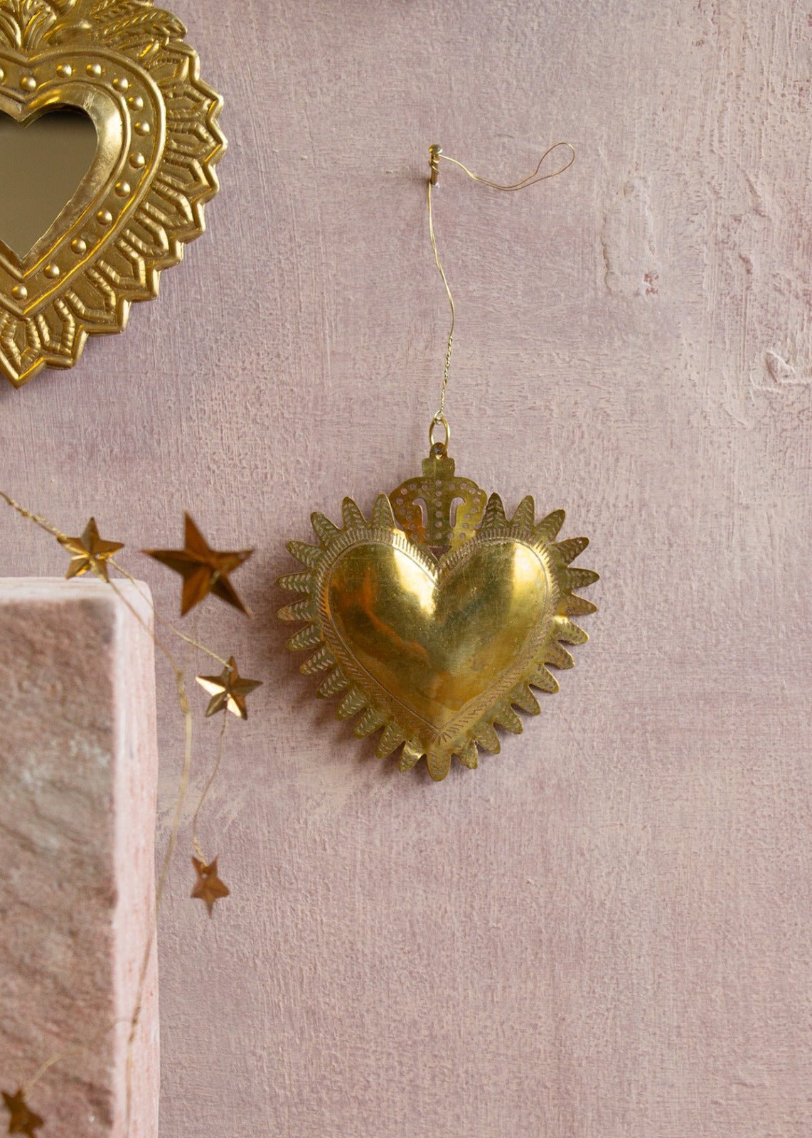 Golden handmade ex-voto heart with embossed crown detail, hanging on a wall with star garland nearby.