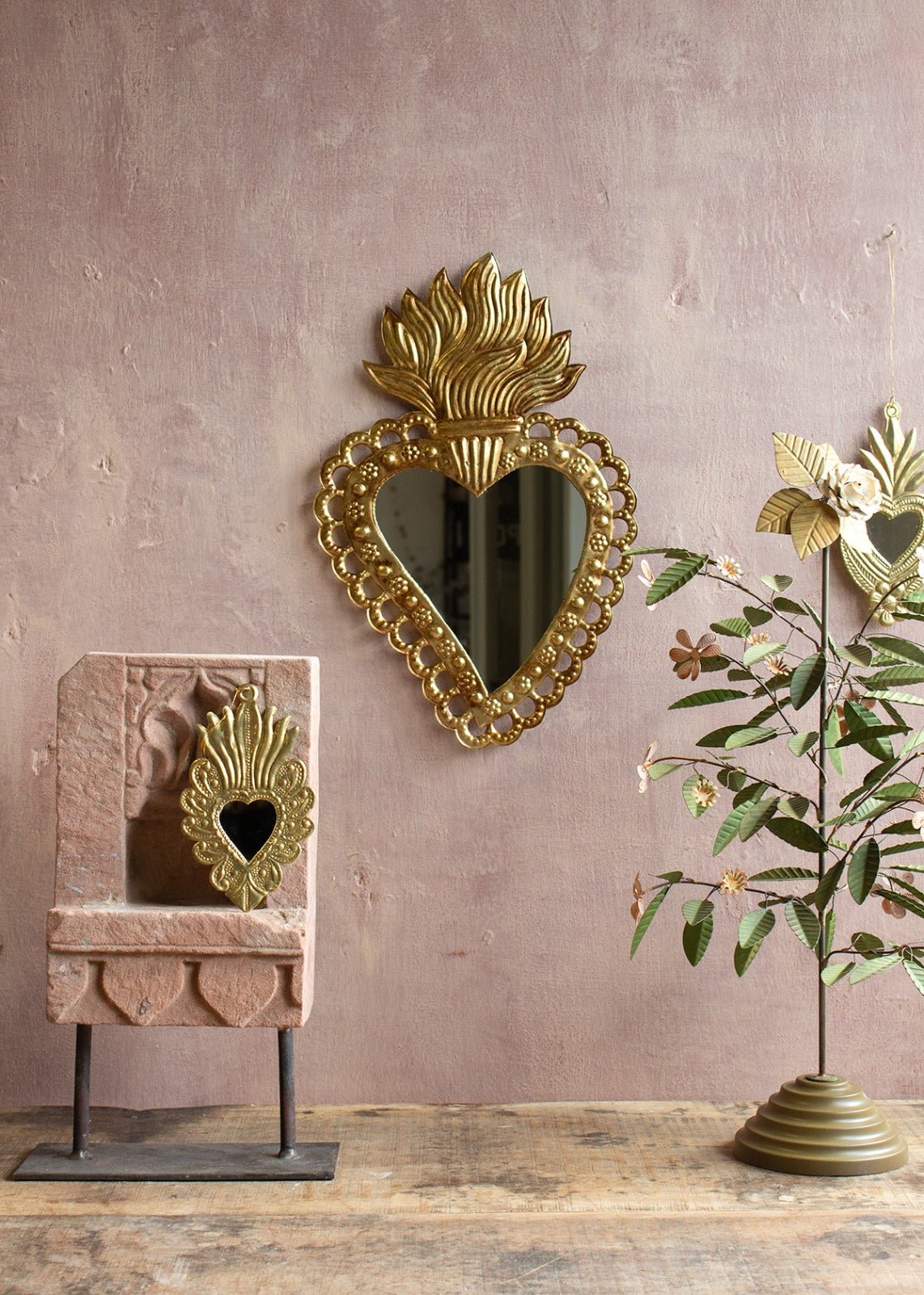 Heart-shaped brass mirror with flame-shaped crown and floral dotted frame, hanging on a pink textured wall with decorative objects and a brass floral tree.