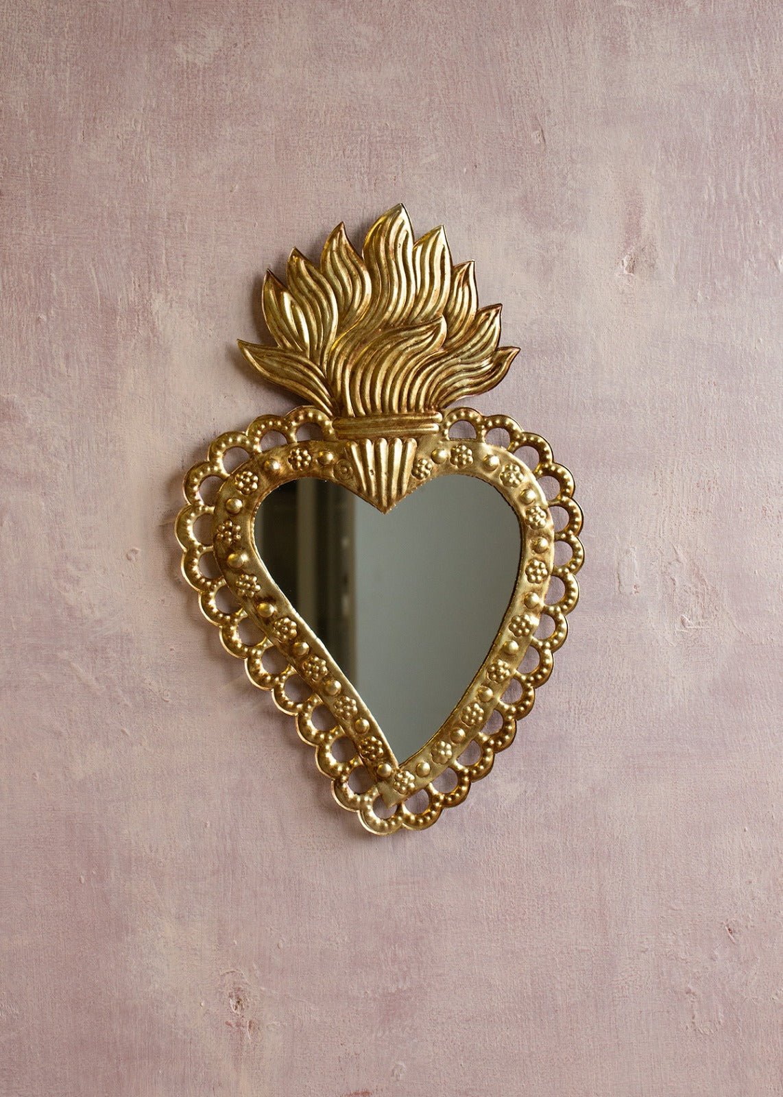 Heart-shaped brass mirror with flame crown and decorative dotted border, hanging on a textured pale pink wall.