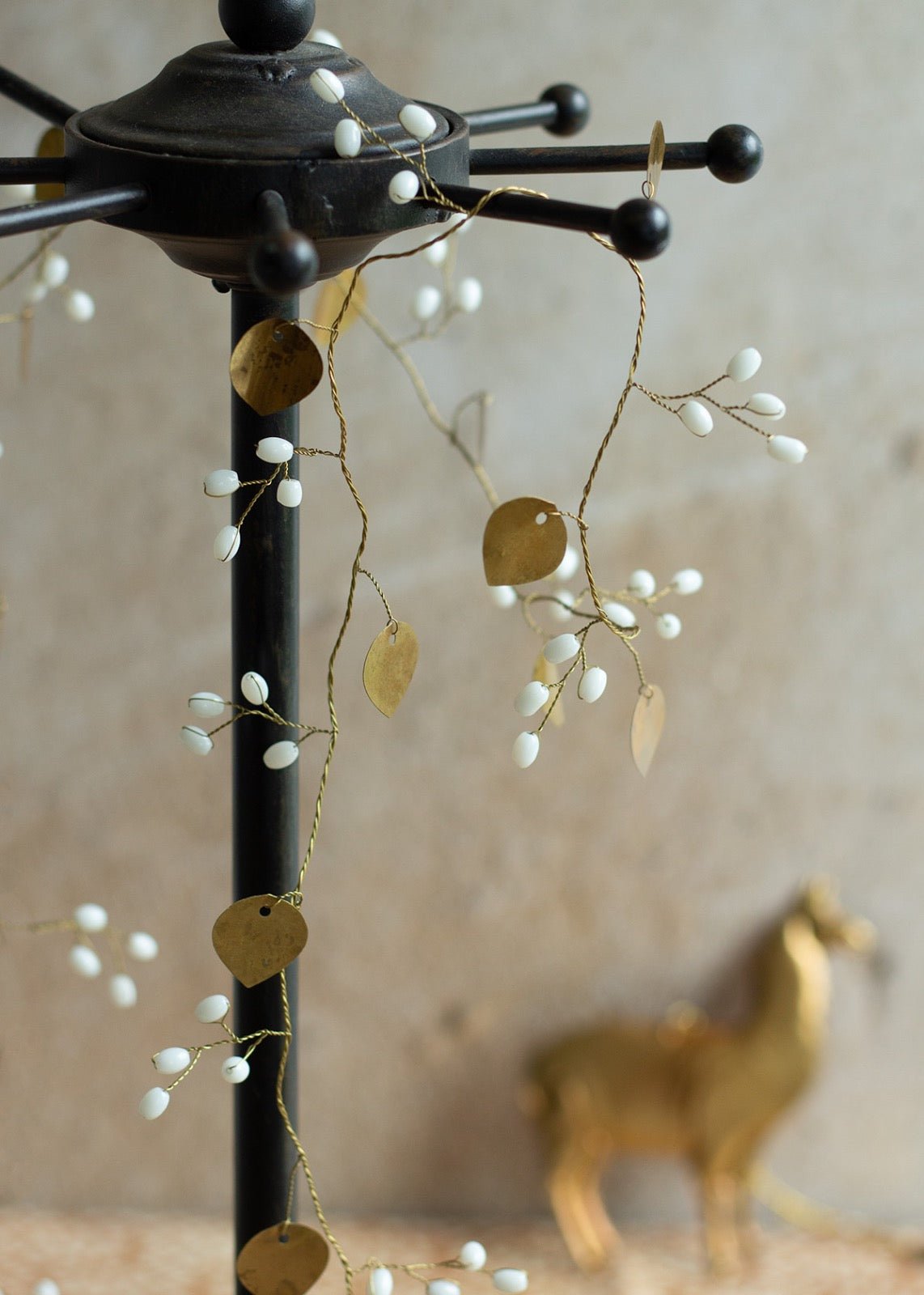 Close-up of a decorative garland with gold heart-shaped metal leaves and small white berry-like beads, wrapped around a black stand with a soft beige background and a golden animal figurine in the distance.