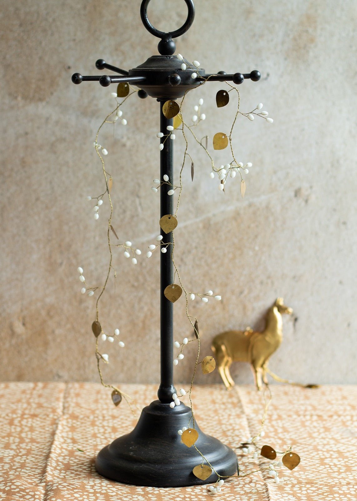 Black metal jewelry stand with a decorative garland of gold heart-shaped leaves and white beads, placed on a patterned cloth with a golden deer figurine in the background