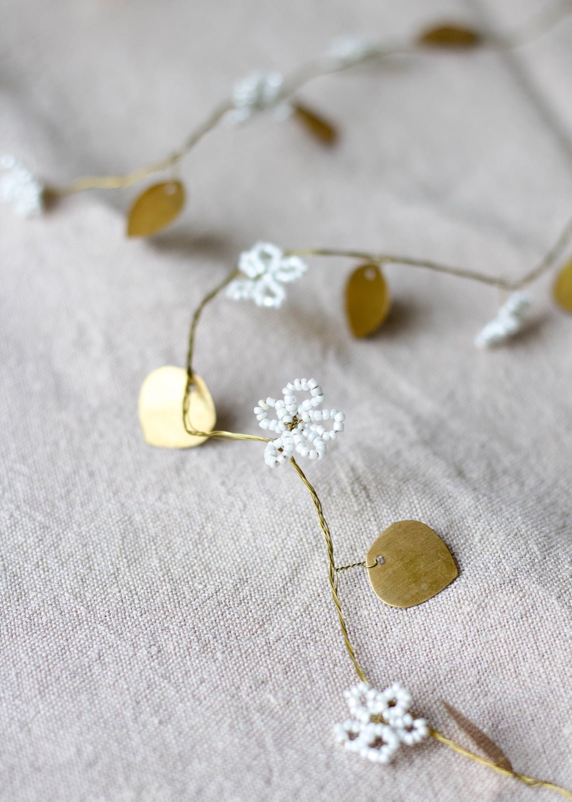 Close-up of a garland featuring white beaded flower blossoms and small gold brass leaves, arranged on a soft beige linen fabric.