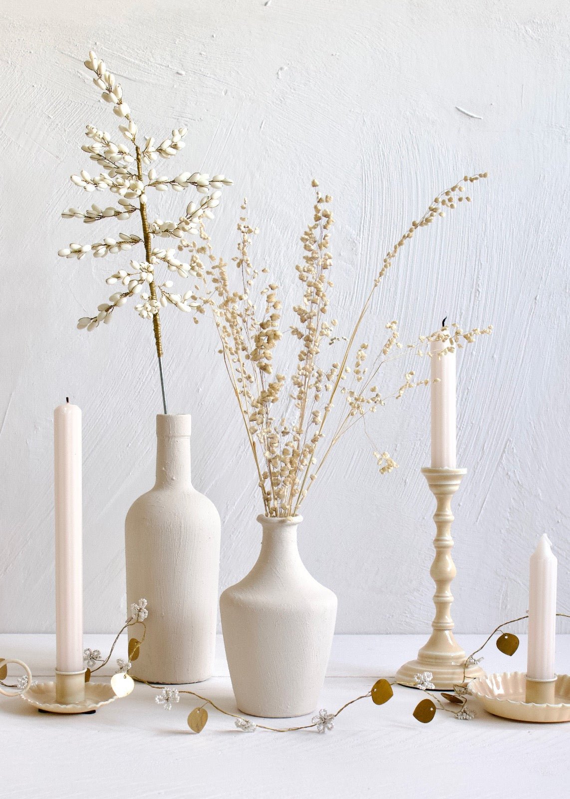 Delicate garland with white beaded flowers and brass leaves arranged across a white surface, styled with cream-toned vases, dried flowers, and tall candles in matching candleholders against a textured white wall.