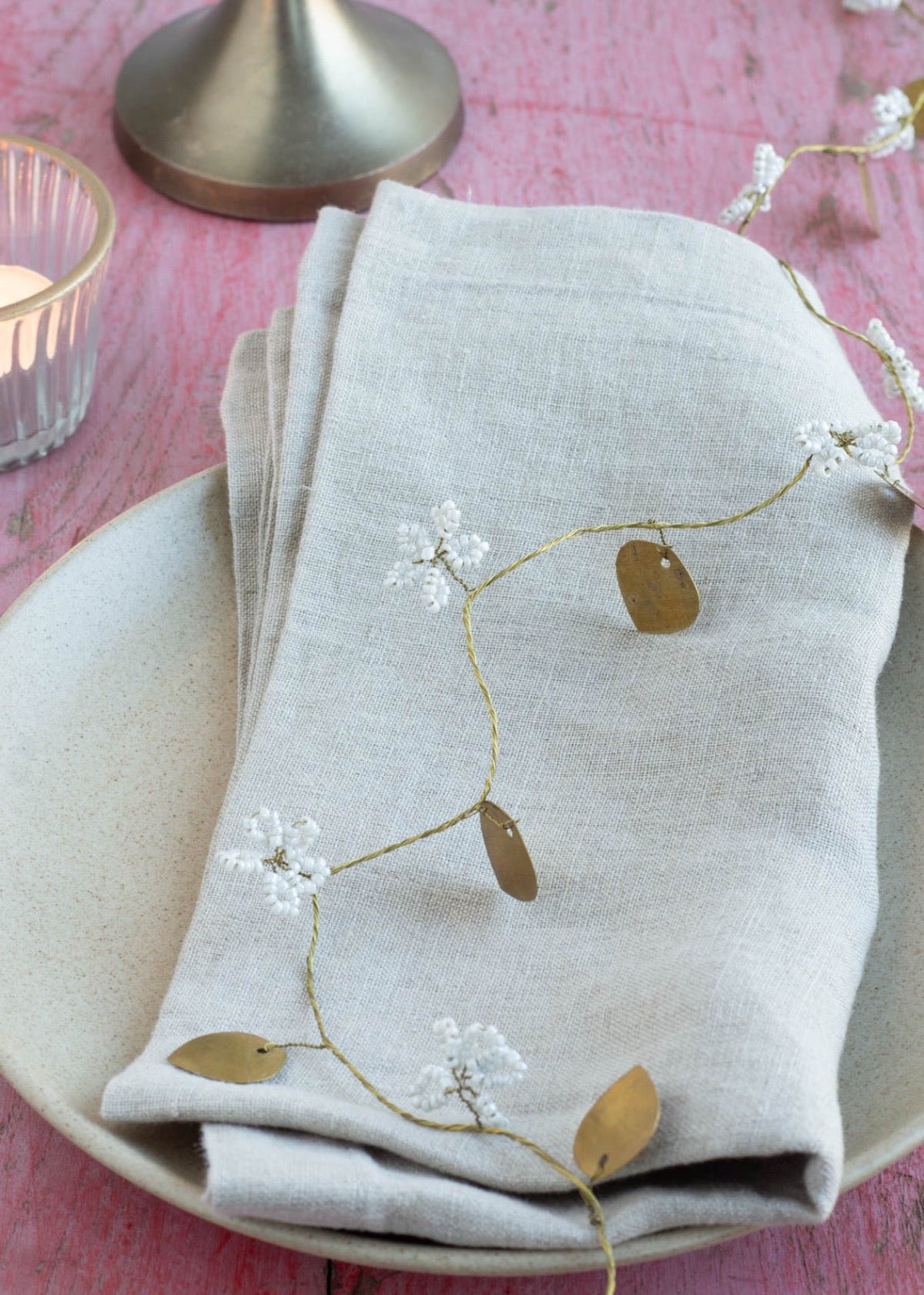 Delicate garland with white beaded flowers and golden brass leaves placed over a folded linen napkin on a ceramic plate, with a candle and pink wooden table in the background.