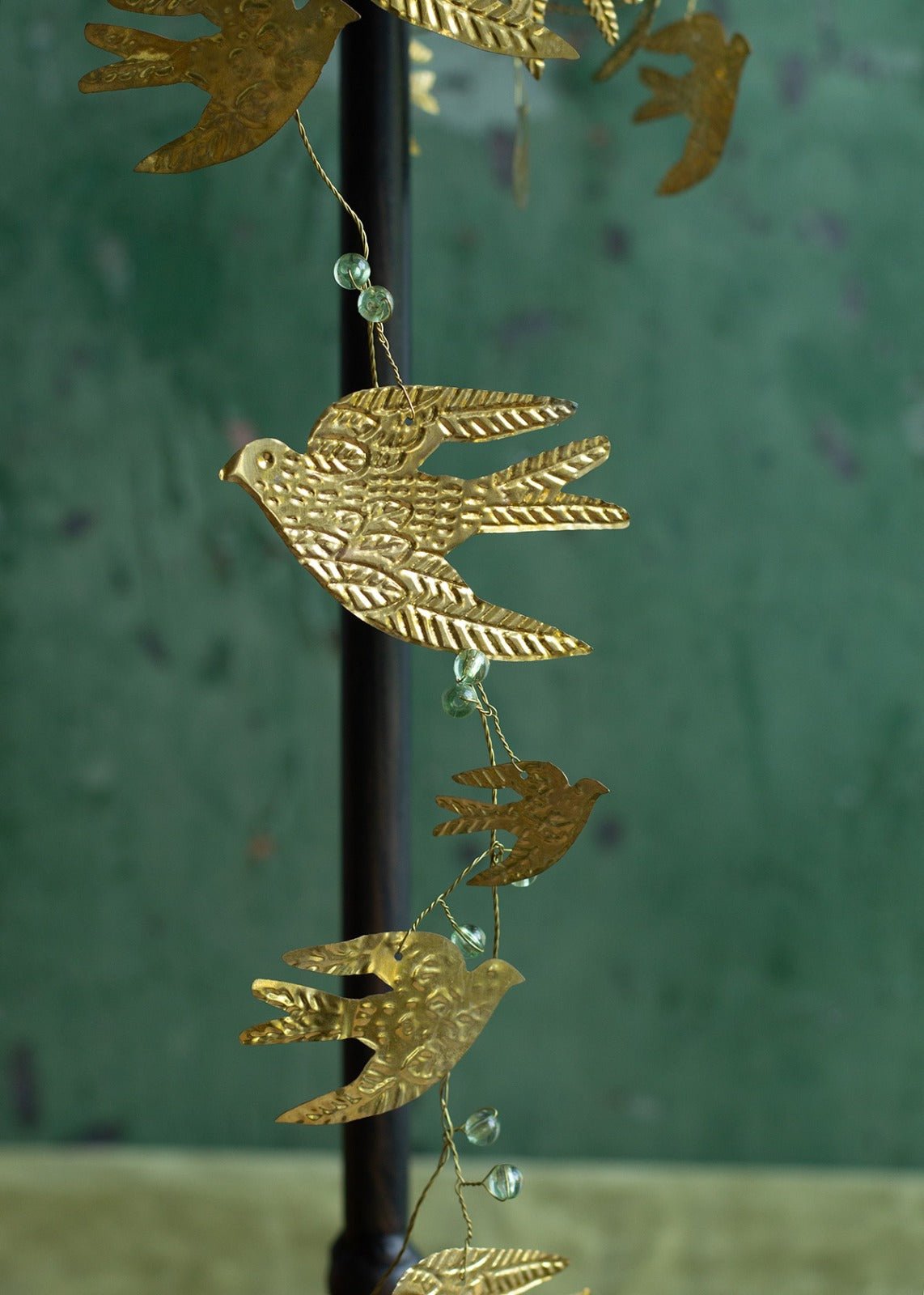 Close-up of a decorative garland with detailed golden brass birds and translucent green glass beads, hanging against a deep green textured wall.