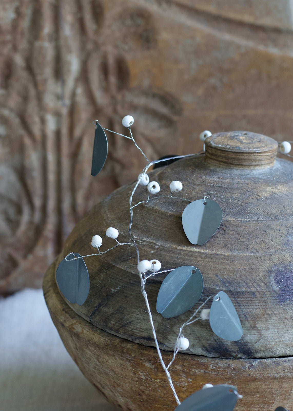 Garland with matte zinc leaves and white beads, draped over a rustic wooden pot with a textured background