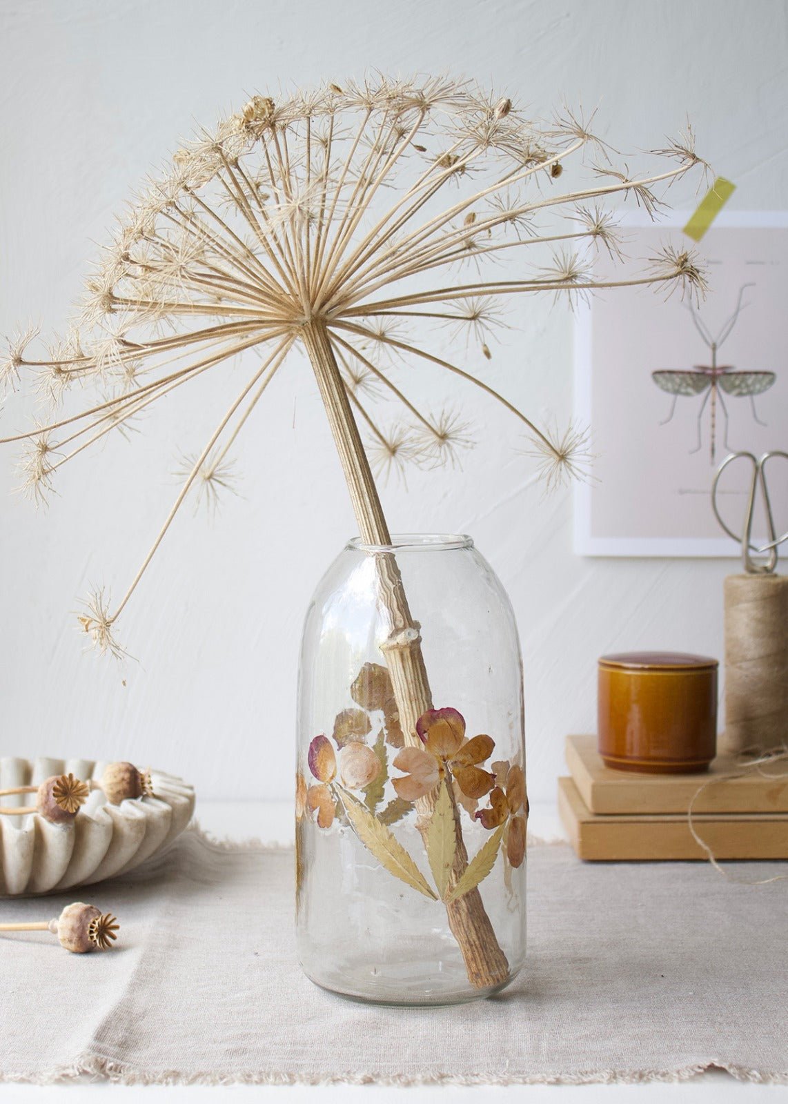 Clear glass vase with real pressed flowers and a dried allium stem on a linen-covered table.