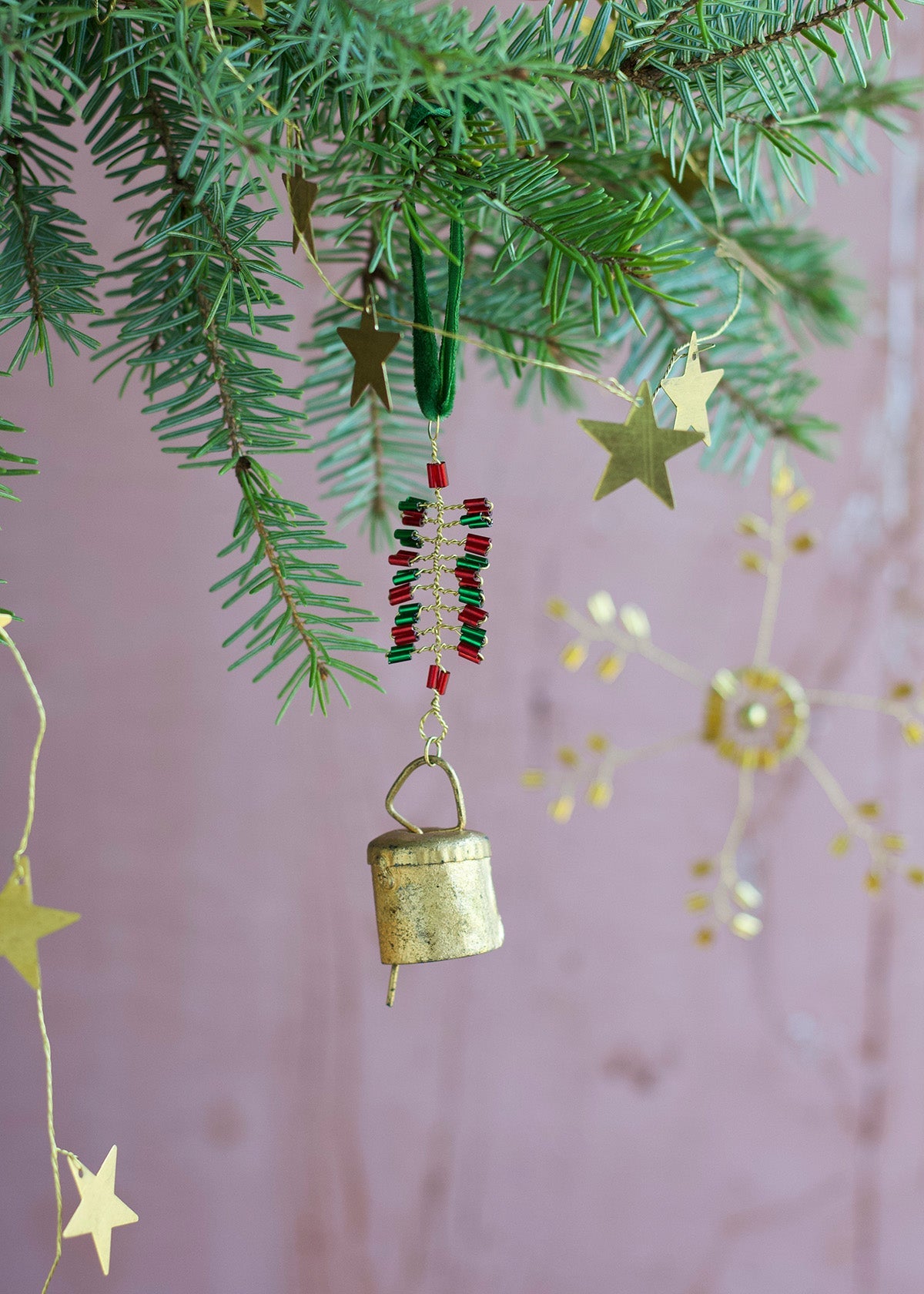 Decorative gold bell and star-shaped ornaments hanging from a Christmas tree branch against a pink background.