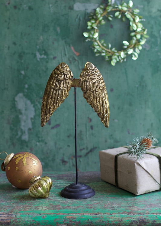 Golden angel wings decor piece on black stand with Christmas ornaments and gift box in the background.