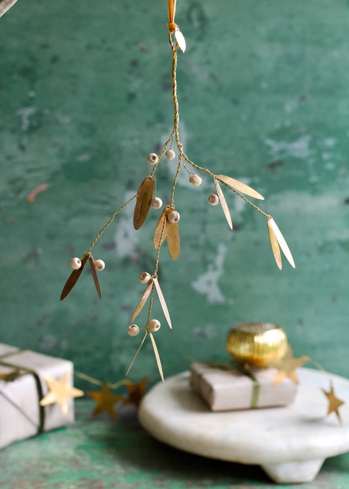 Golden mistletoe ornament with pearl-like berries hanging in front of green background with wrapped presents.