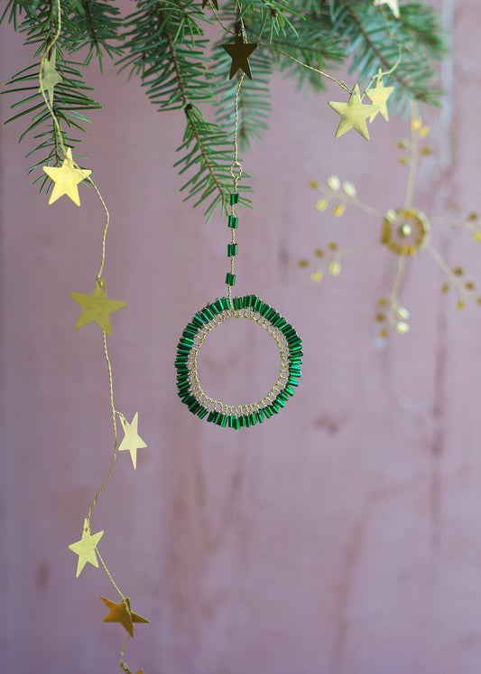 Decorative green beaded circle ornament with gold stars hanging from a branch against a soft pink background.
