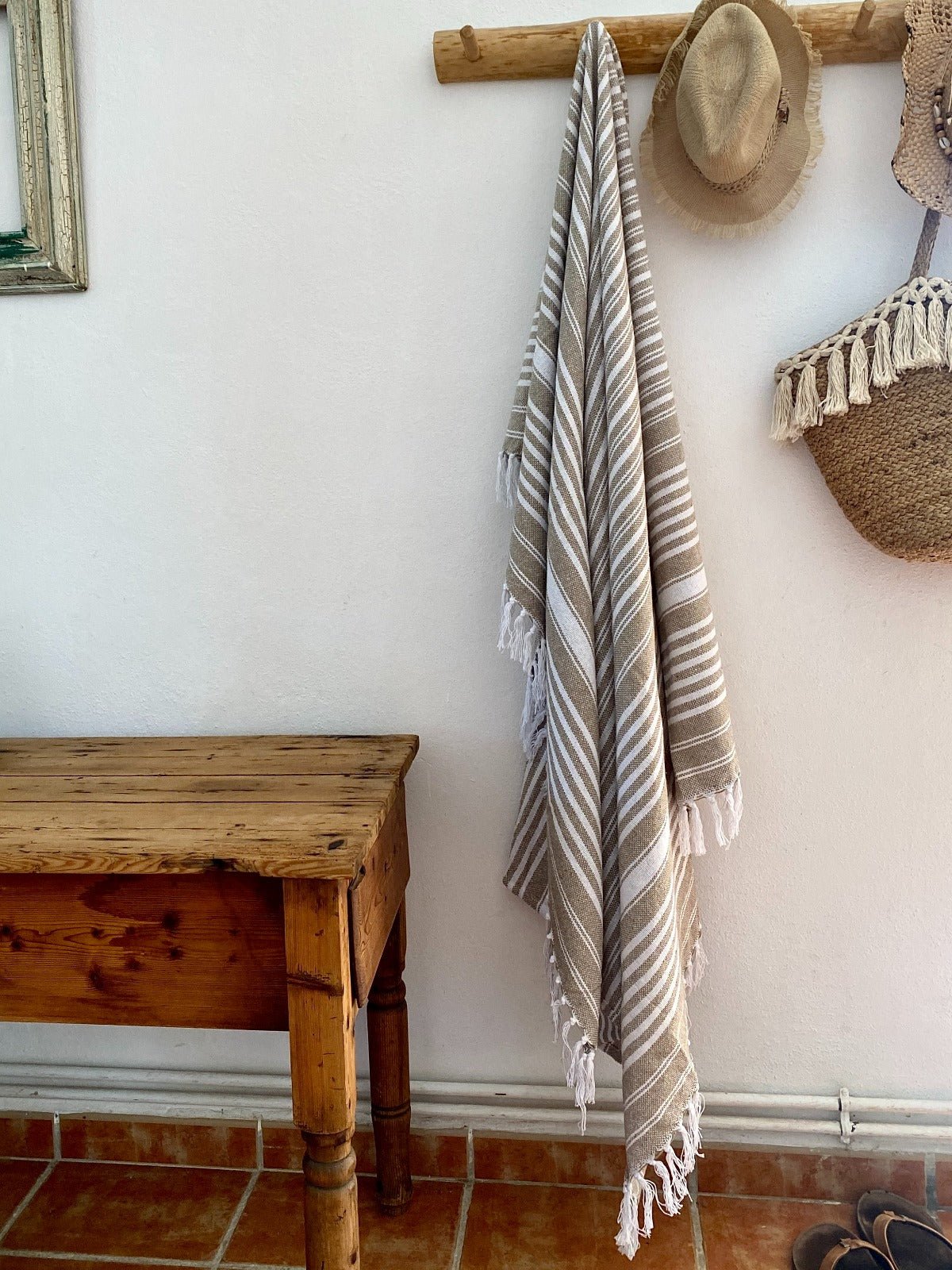 Towel with tassels hanging on a wall next to a wooden bench and hats.
