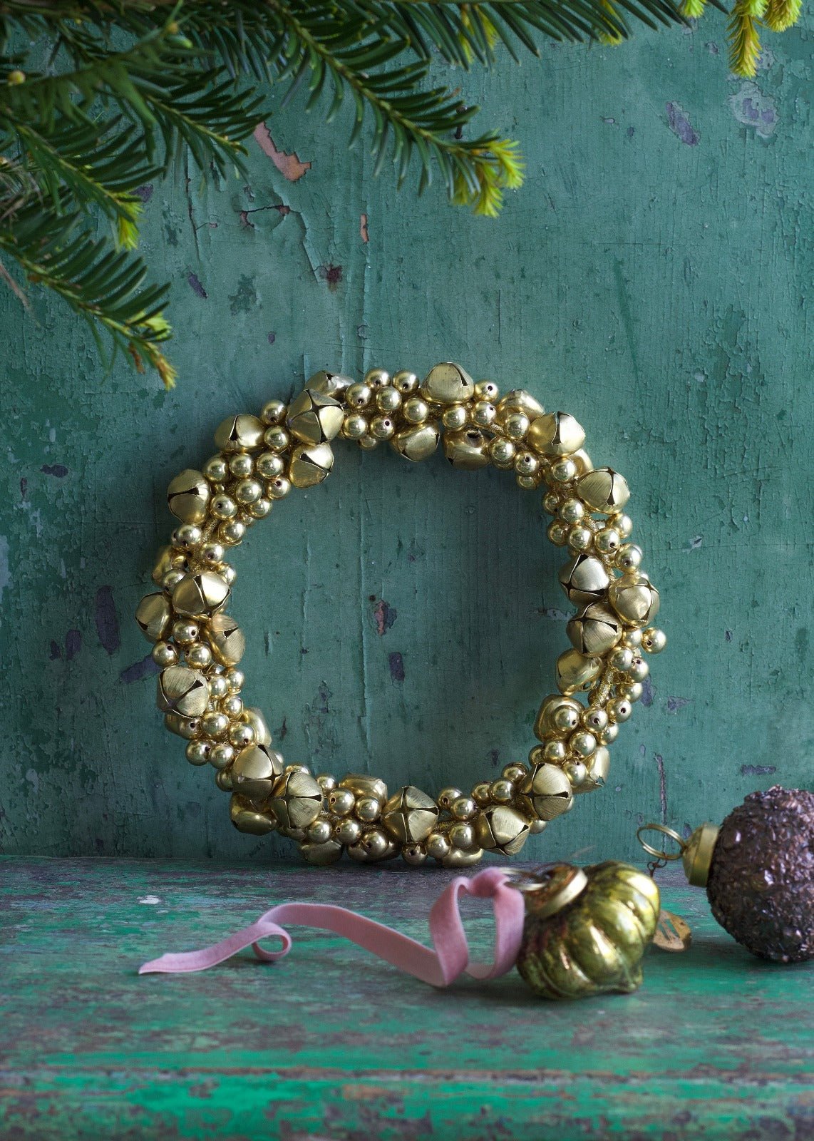 Round handmade wreath made of many small golden bells, styled against a rustic green background with Christmas ornaments nearby.