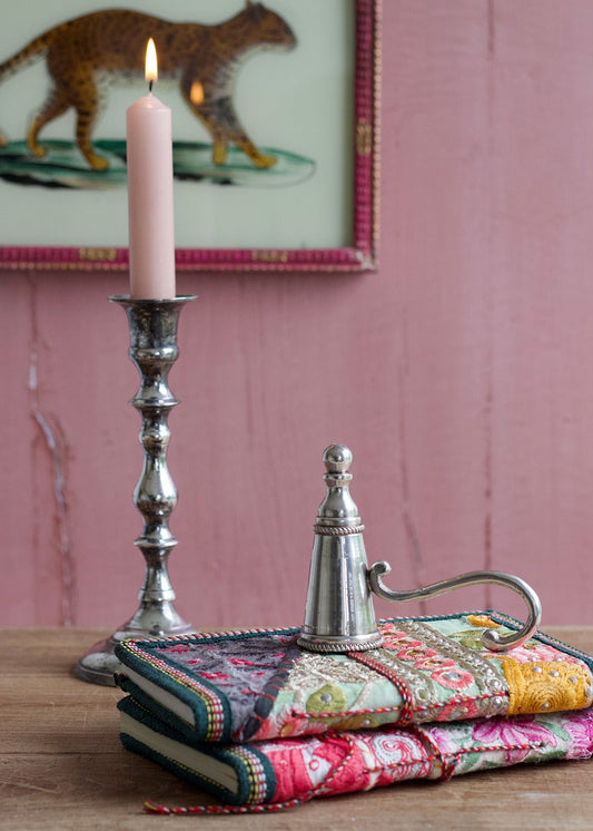 Decorative stack of books with a candle and silver container on a wooden surface, with a pink wall and framed picture in the background.