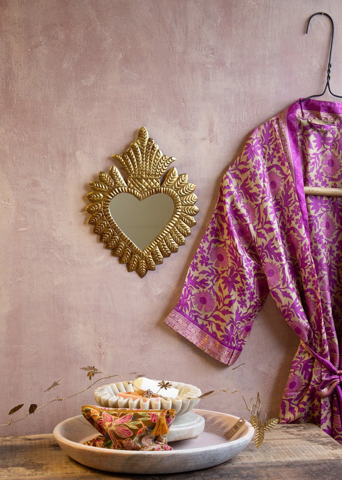 Heart-shaped brass mirror with embossed rays and crown detail, hanging on a blush pink wall next to a purple and gold floral kimono and marble accessories with golden garland.