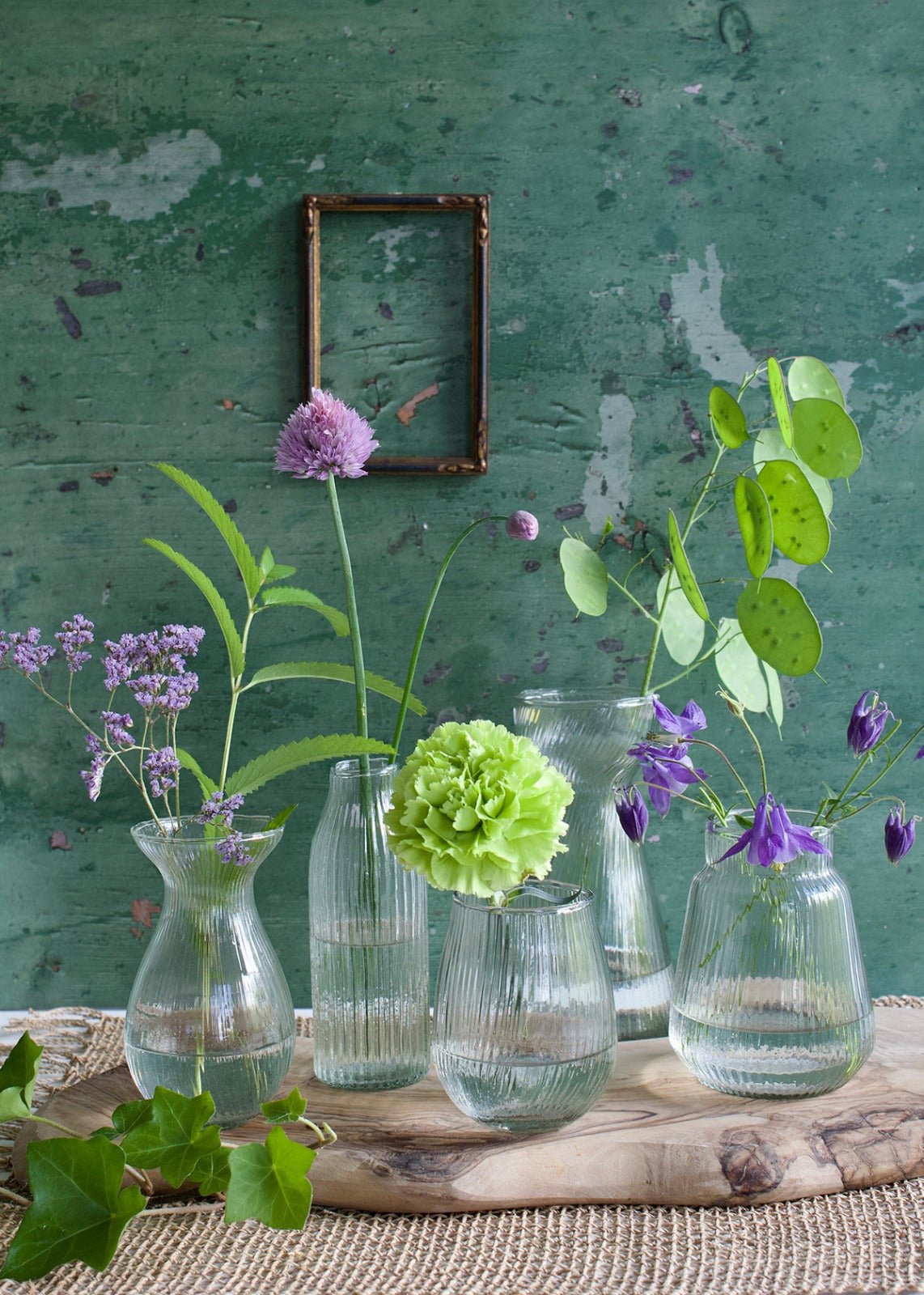 Collection of small ribbed glass vases in various shapes, each holding spring flowers, arranged on a rustic wooden board with a distressed green wall in the background.
