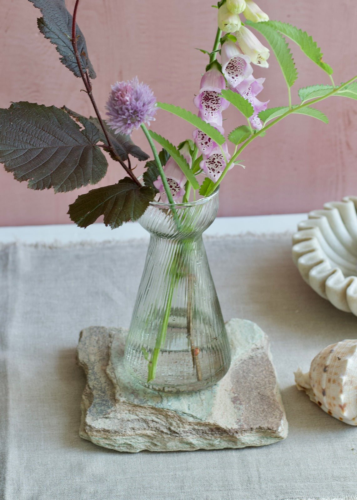 A ribbed glass hyacinth vase with a flared neck, filled with wildflowers and placed on a textured stone slab. The soft pink background and natural linen tablecloth create a serene, elegant setting with decorative elements like a ceramic bowl and a seashell nearby.