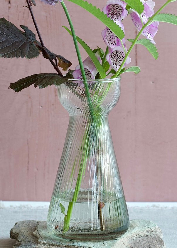 Slender ribbed glass vase with a pinched neck and flared base, styled with soft lilac and green wildflowers.
