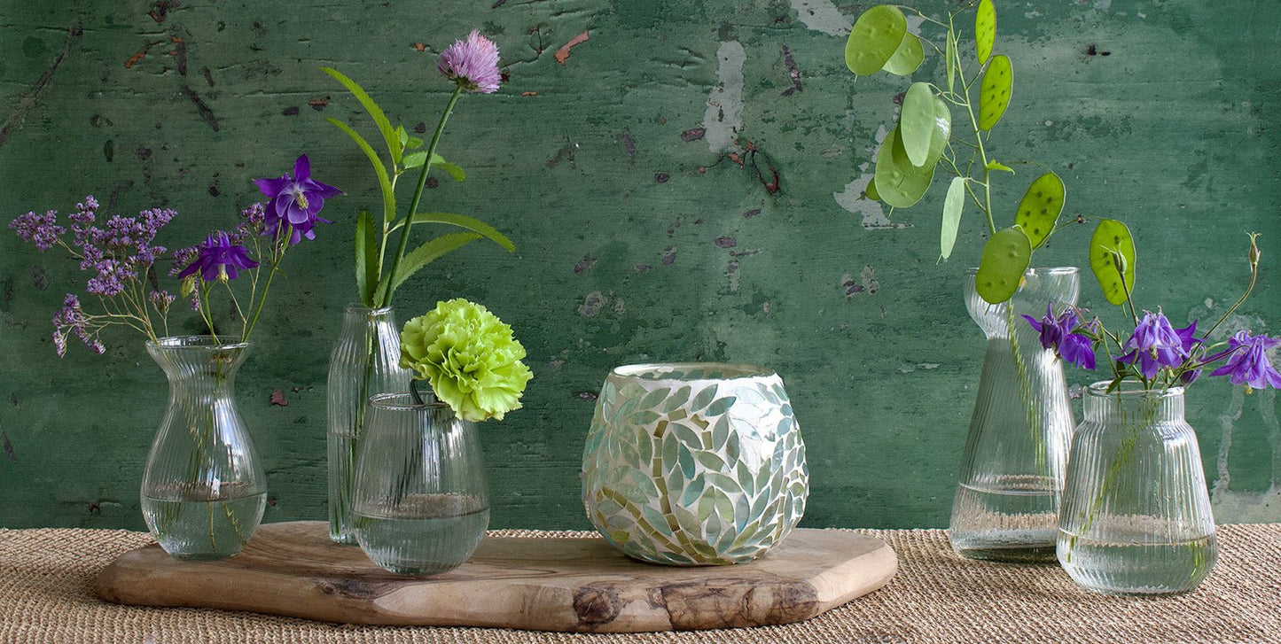 A collection of ribbed glass vases in various shapes filled with fresh flowers, arranged on a wooden tray alongside a round mosaic tealight holder. The setting features a rustic green wall in the background and a woven placemat underneath.