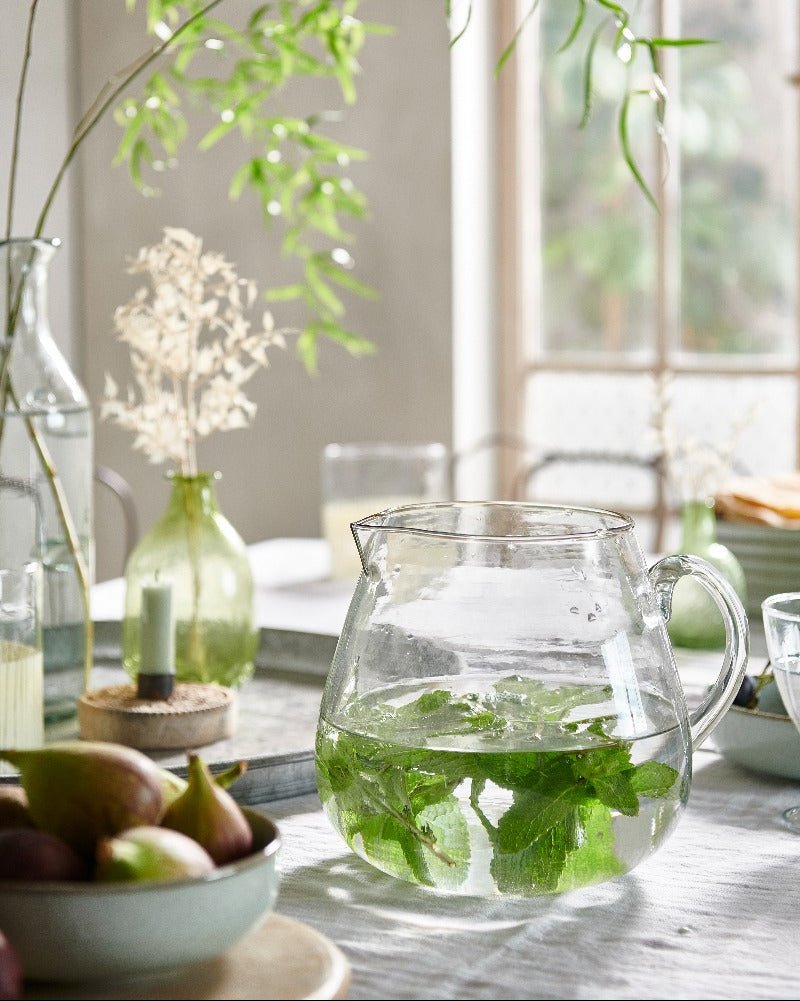 Clear glass pitcher with water and mint leaves on a table with a blurred indoor background