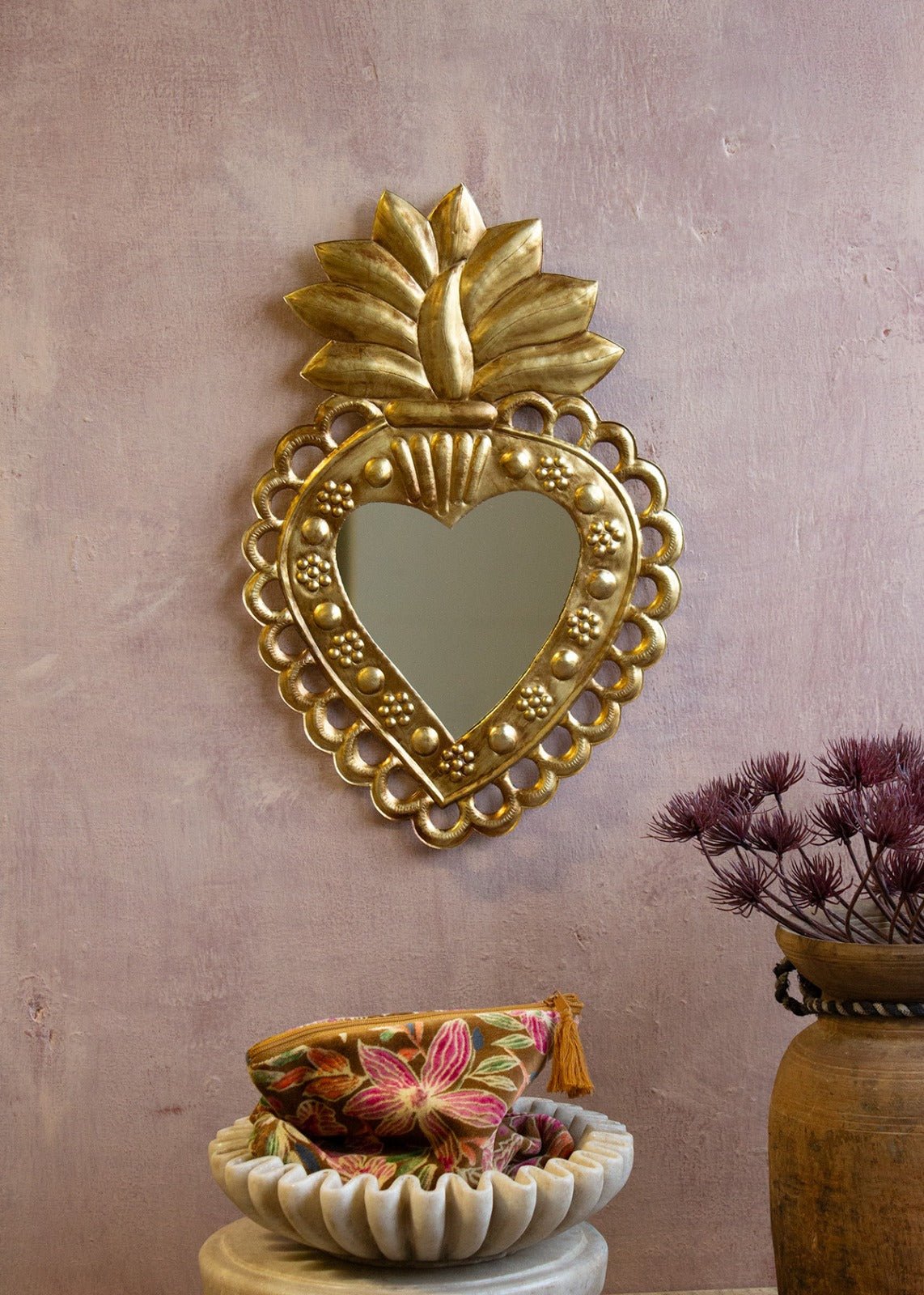 Heart-shaped brass mirror with floral dotted details and a radiant crown on top, hanging on a pink textured wall next to a velvet pouch and dried flowers.