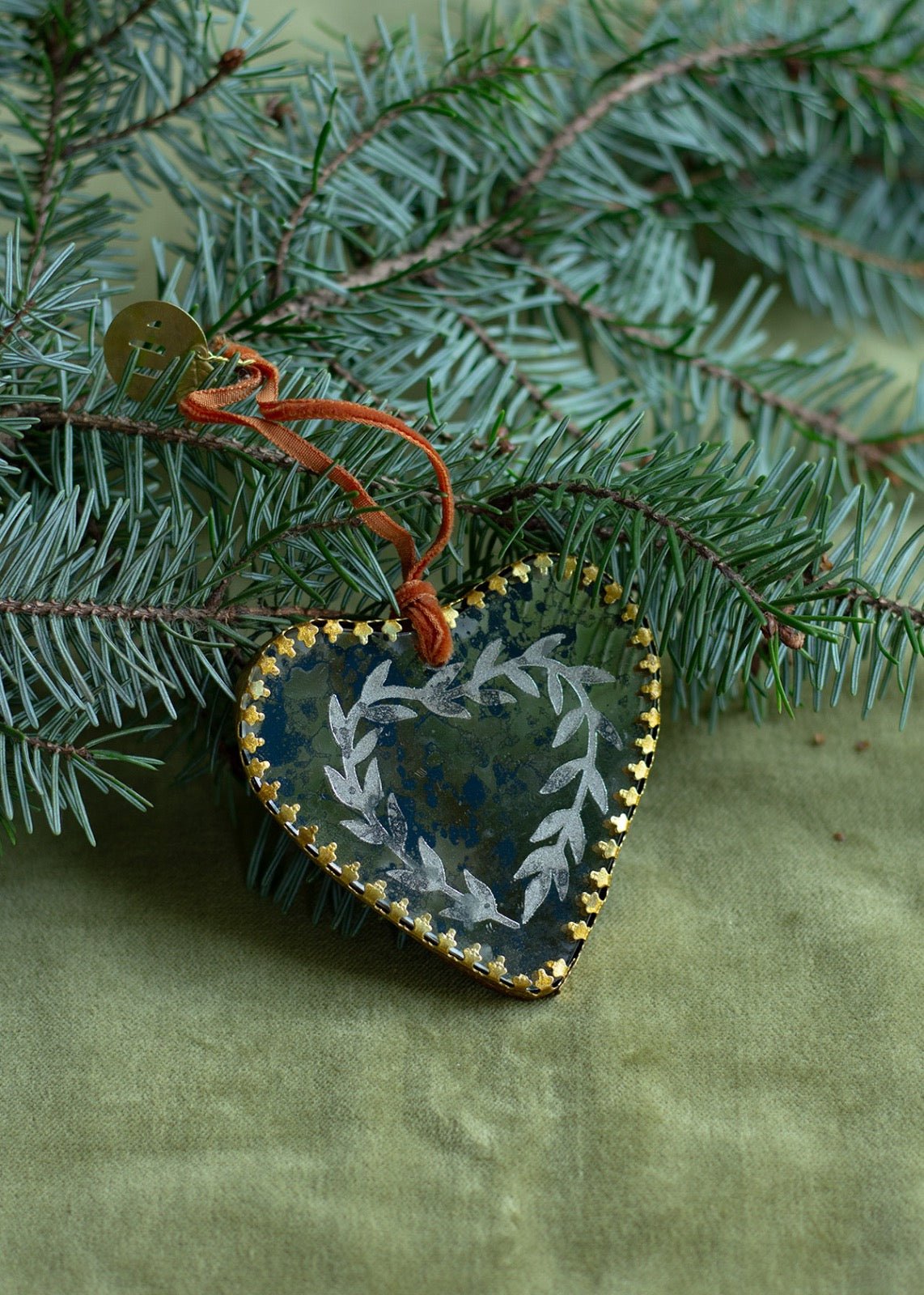 Dark antique-style glass heart ornament with golden edges and leaf details, lying on a fir branch.