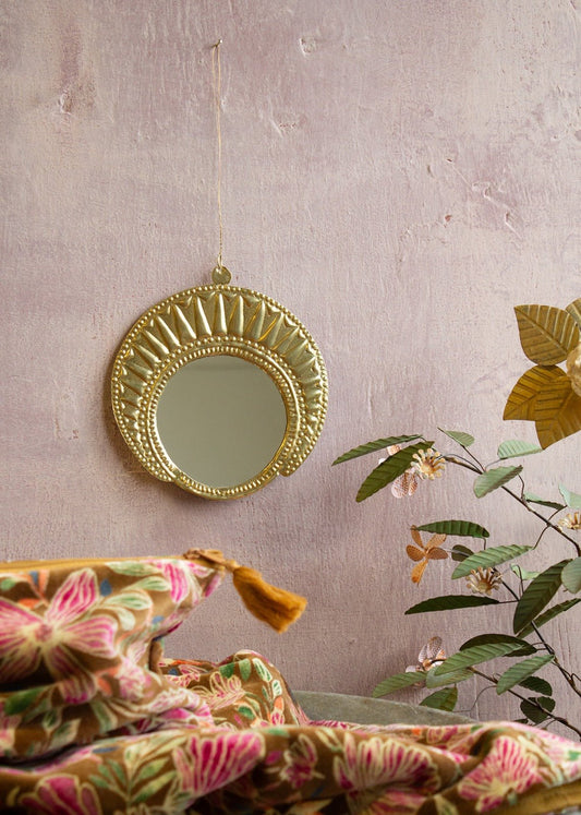 Round gold-toned metal wall mirror with radiant embossed detailing, handcrafted and hung against a soft pink wall.