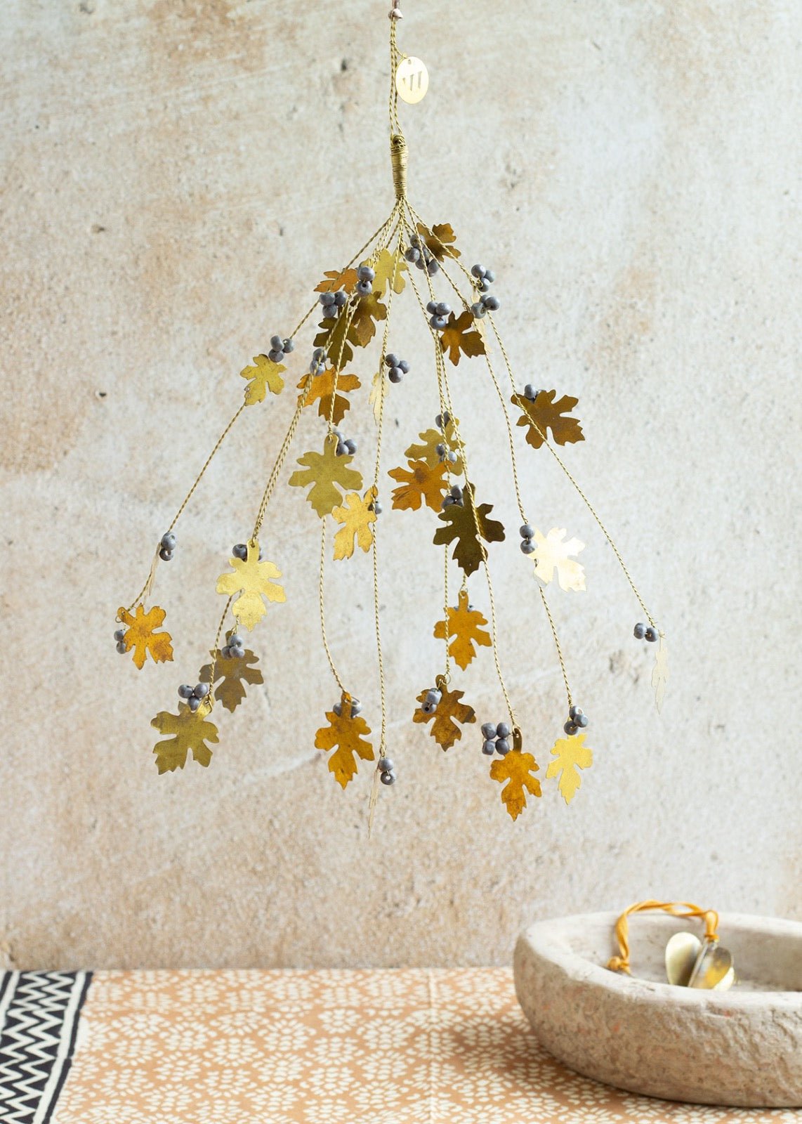 Decorative brass bunch with oak leaf shapes and grey beads, hanging against a textured beige wall above a patterned surface with a stone bowl.