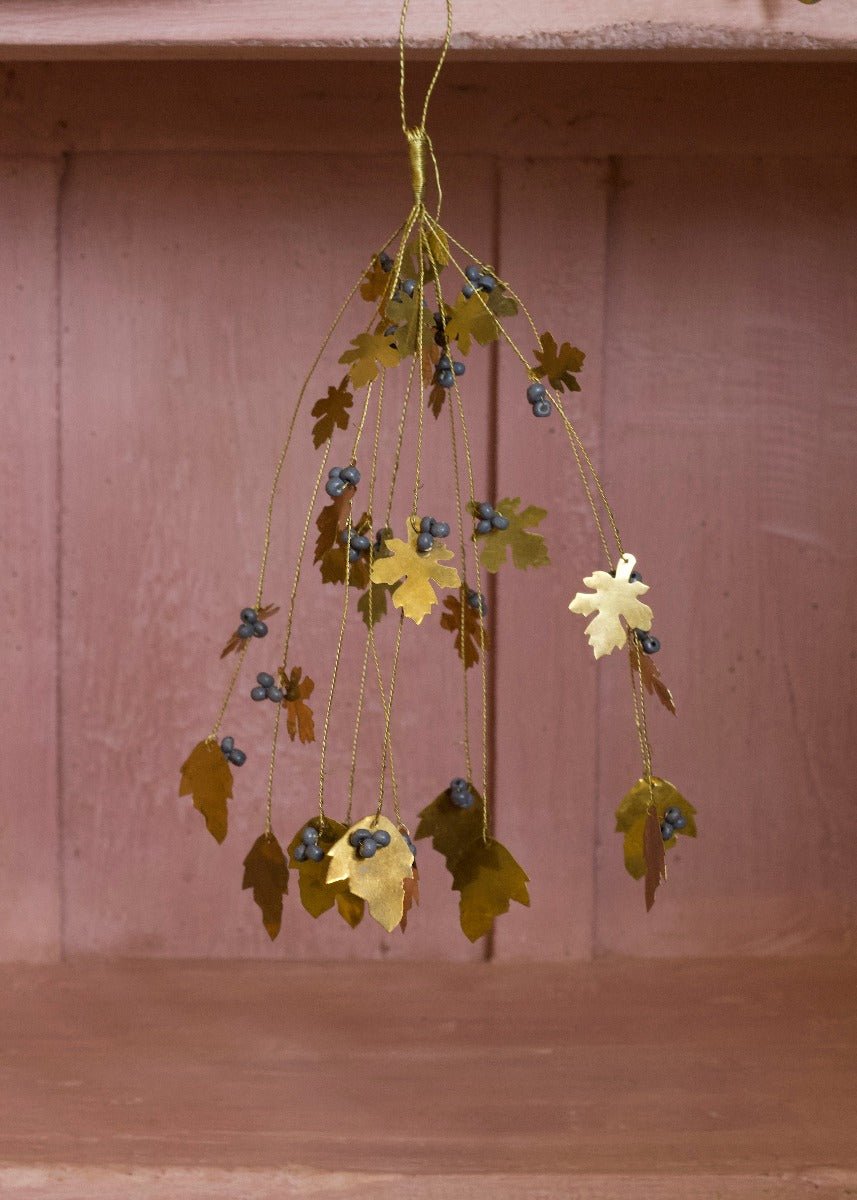 Hanging brass bunch with oak-shaped leaves and grey beads, displayed against a soft pink wooden background.