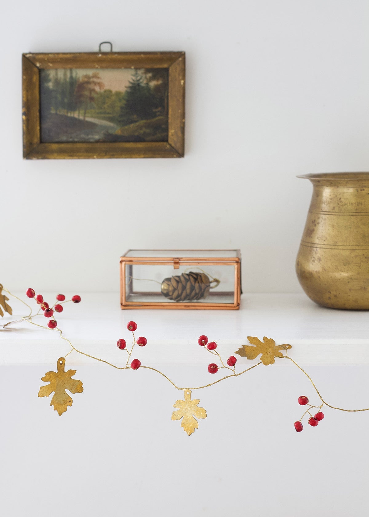 Decorative gold leaf and red berry garland on a white surface with a framed picture and brass container in the background.