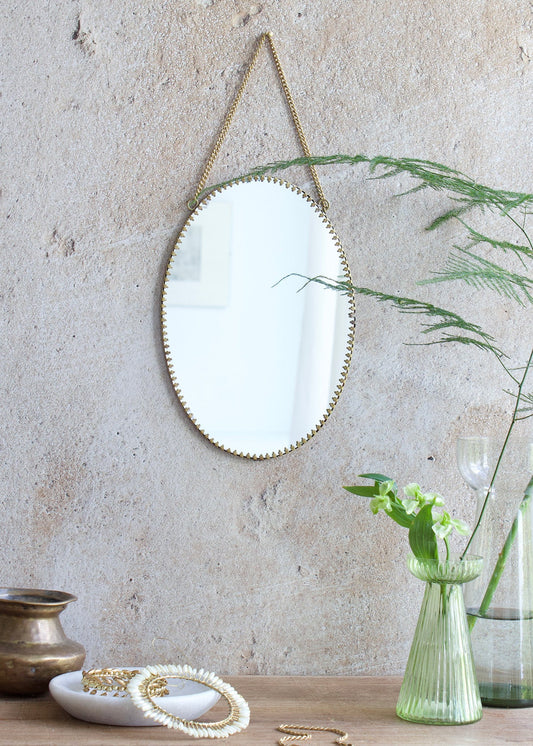 Oval mirror with a decorative brass edge, hanging from a brass chain against a textured wall, styled with green flowers, vintage glassware, and golden accessories.